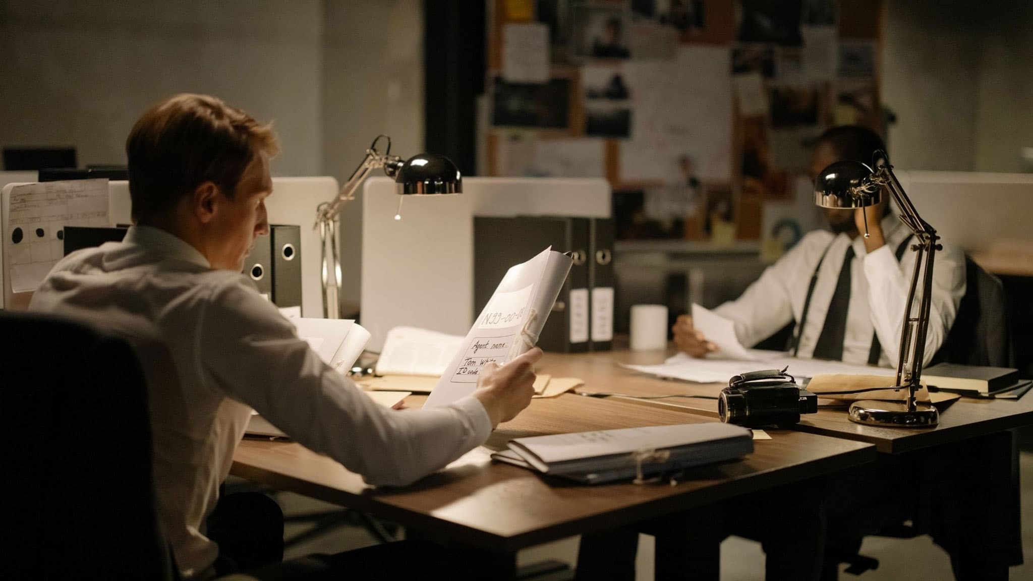 Two investigators reviewing case files at desks in a dimly lit office, with an evidence board of photographs and notes on the wall behind them